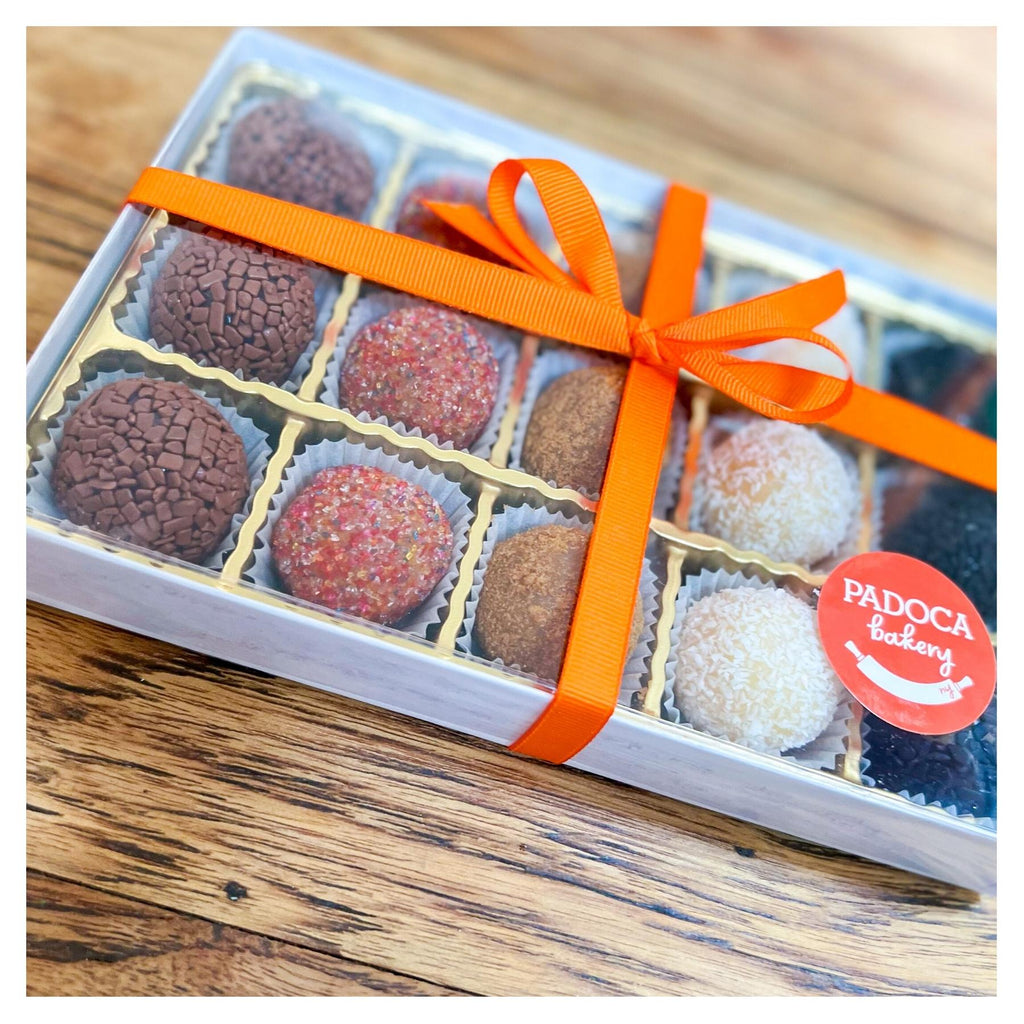 A box 15 mixed flavors of Brazilian Brigaderios - chocolate truffles.  The box has a clear top and is wrapped with an orange bow and finished with an circle orange Padoca Bakery sticker. 