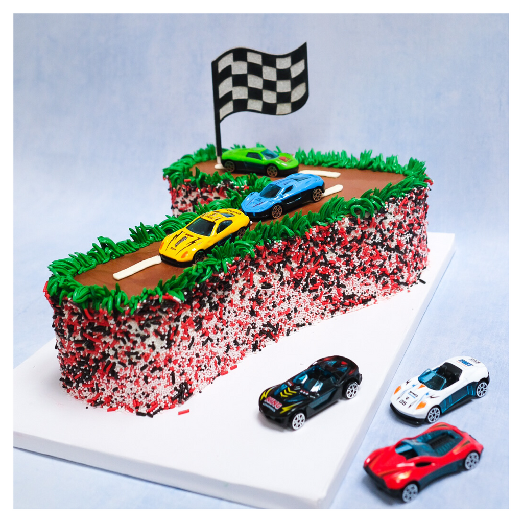 A picture of a number 7 shaped cake decorated in a race car theme.  The top is a brown and white race track with three race cars, green, yellow and blue.  Also on top of the cake is a race car flag and green piping to mimic grass.  The sides of the cake are a mixture of red, black and white sprinkles.  Next to the cake board are three additional cars - the colors are black, white and red. 
