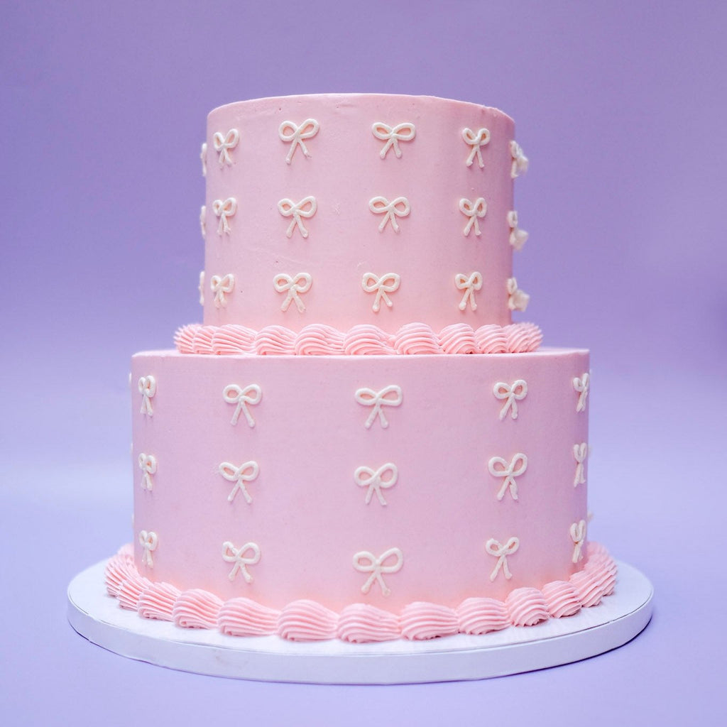 A  two tiered pink cake with pink piping and white ribbon. Slightly different angle