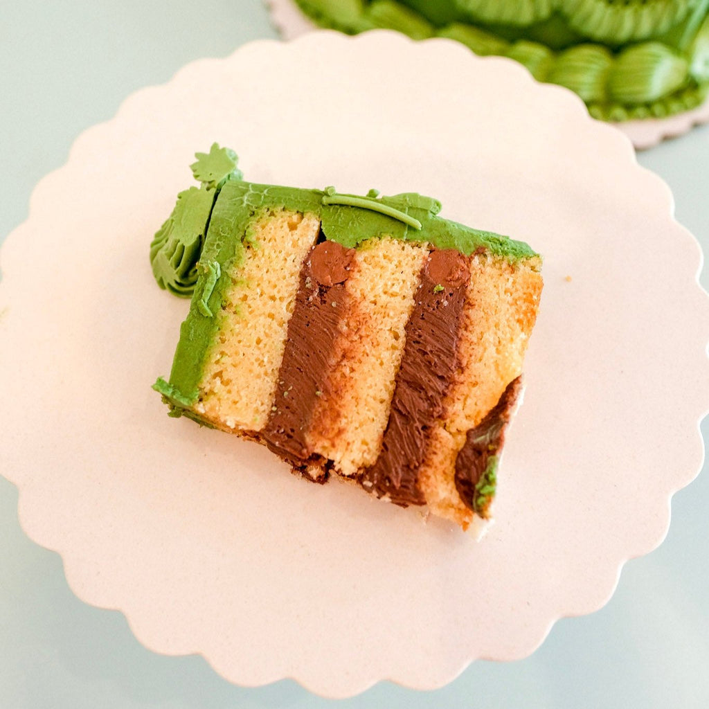 A slice of cake.  Three layers of vanilla cake, two layers of chocolate mousse, topped with green buttercream frosting.  On a white plate. 