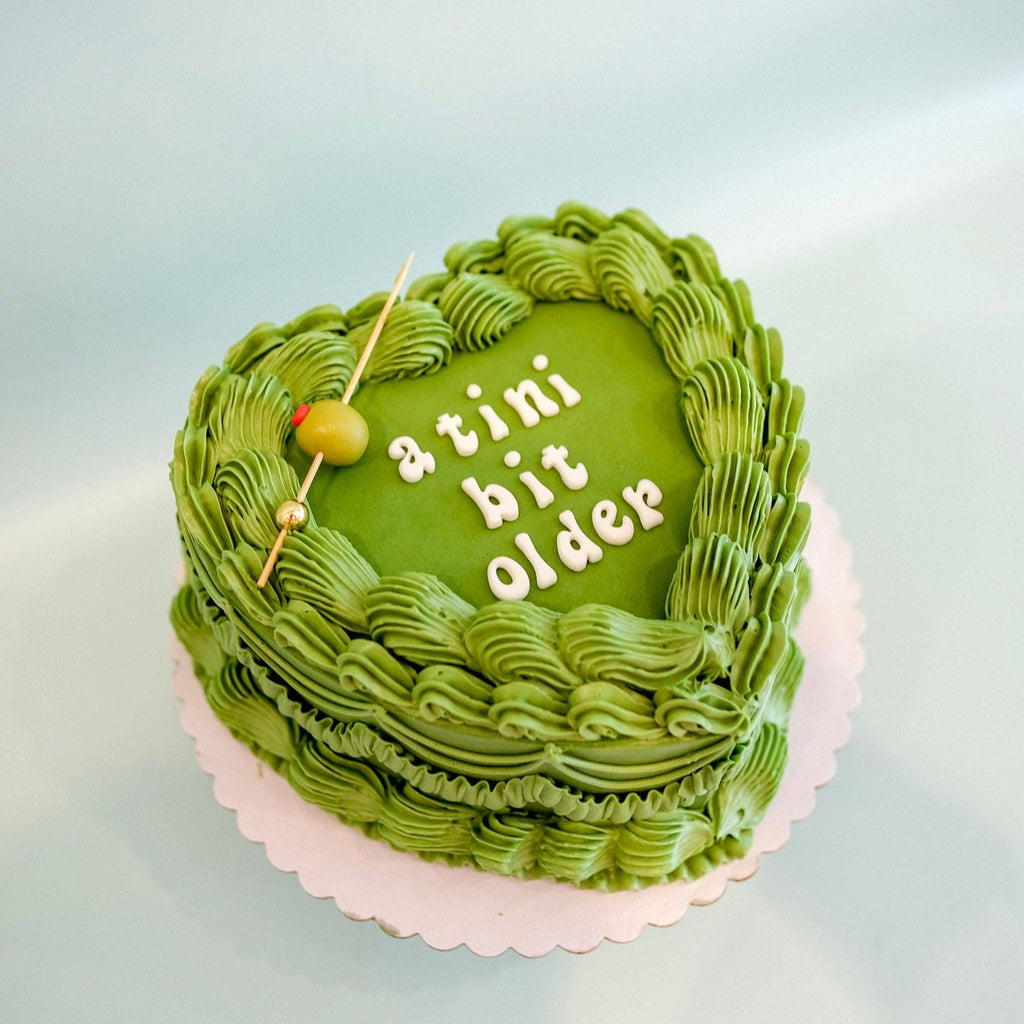 A tini bit older green heart shaped cake.  The cake has green ornate piping a fondant olive with a stick and fondant letters that say a tini bit older. 