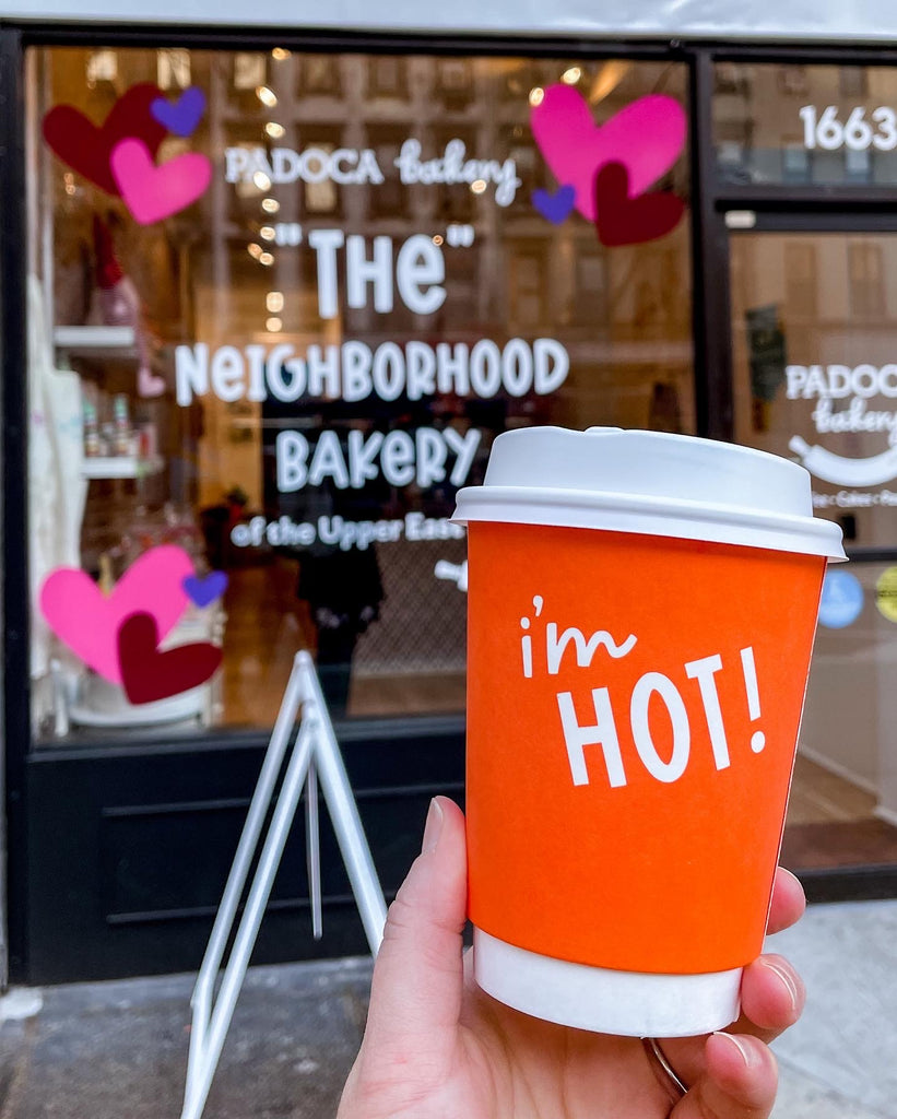 A picture of the exterior of Padoca Bakery.  The window says The Neighoborhood Bakery of the Upper East Side with Valentine's Day hearts in Pink, Maroon and Puple. And a women's hand holding an orange I'm hot coffee mug. 