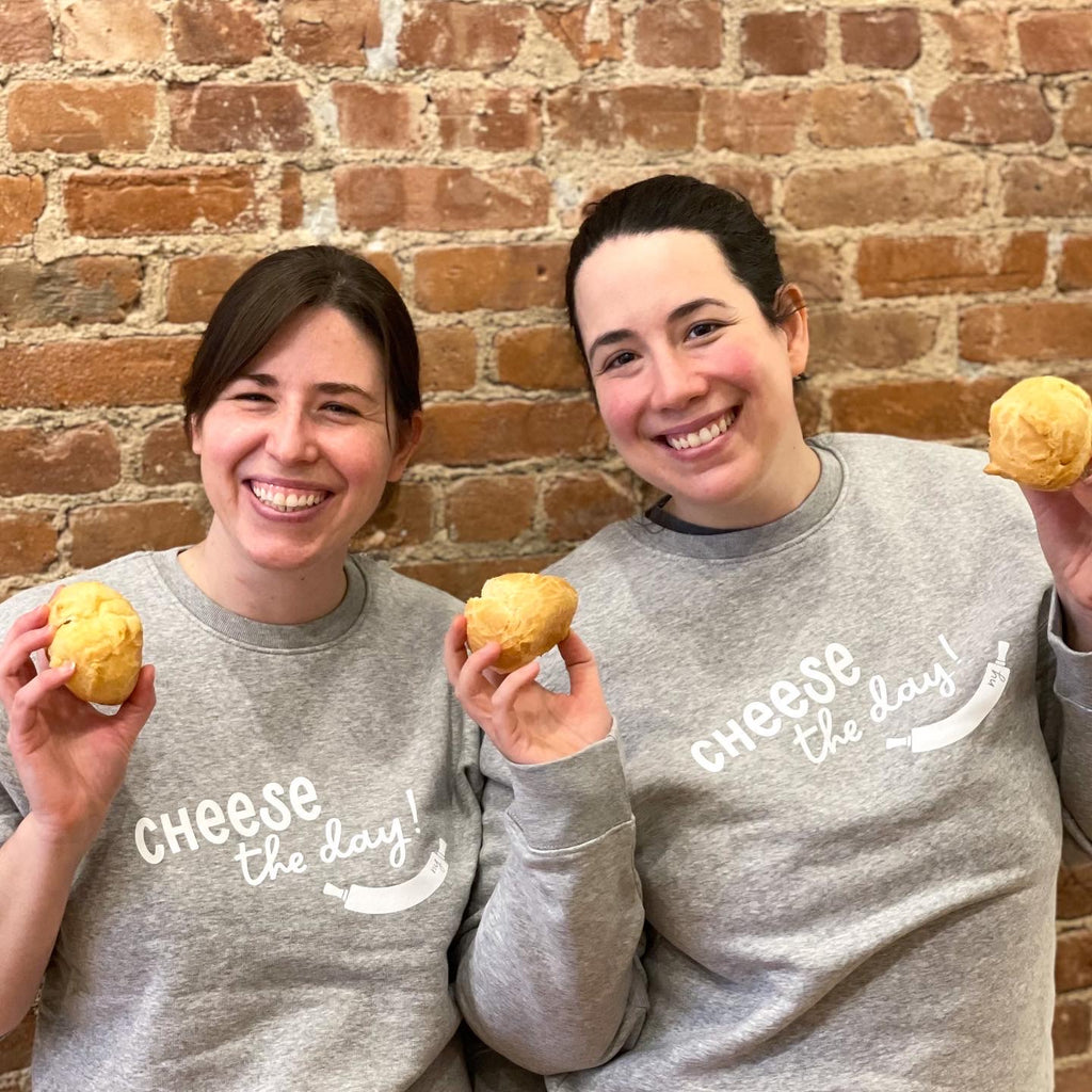 A picture of the Padoca Bakery owners, Marina and Megan smiling together leaning on a brick wall.  They are holding Pao de Quiejos and wearing a grey sweater that says Cheese the Day. 