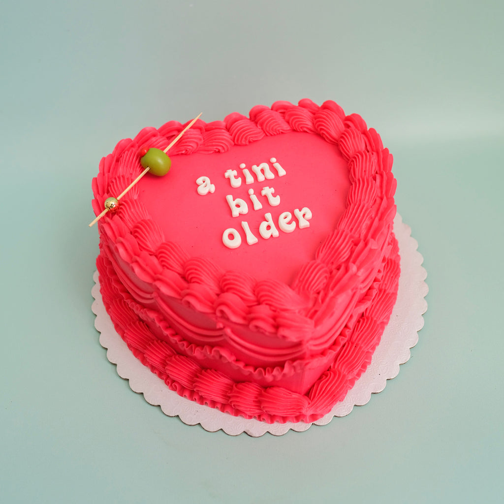 A pink tini bit older heart shaped cake.  The cake has pink ornate piping a fondant olive with a stick and fondant letters that say a tini bit older. 
