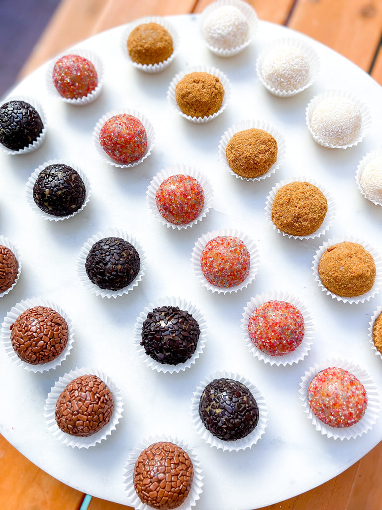 A picture of a mixture of flavors or Brazilian Brigadeiros.  There are 25 Brigadeiros in the photo and they are placed evenly in a row on a white tray. 