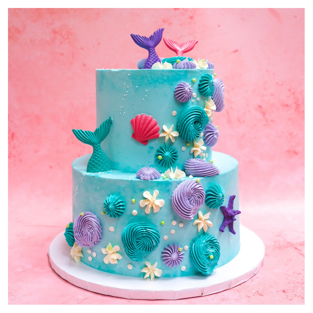 A picture of a mermaid cake.  The cake is two tiered and decorated in a mermaid theme.  Pink, purple and teal candy mermaid fins and sea shells.  The cake is a teal blue with teal blue, purple and white piping and green and white sprinkles. 
