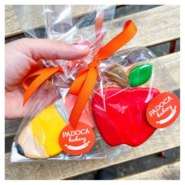 A picture of yellow pencil sugar cookies and red apple sugar cookies wrapped in a clear bag, tied with an orange ribbon and finished with an orange Padoca Bakery sticker. A woman's hands with light pink nail polish is holding them. 